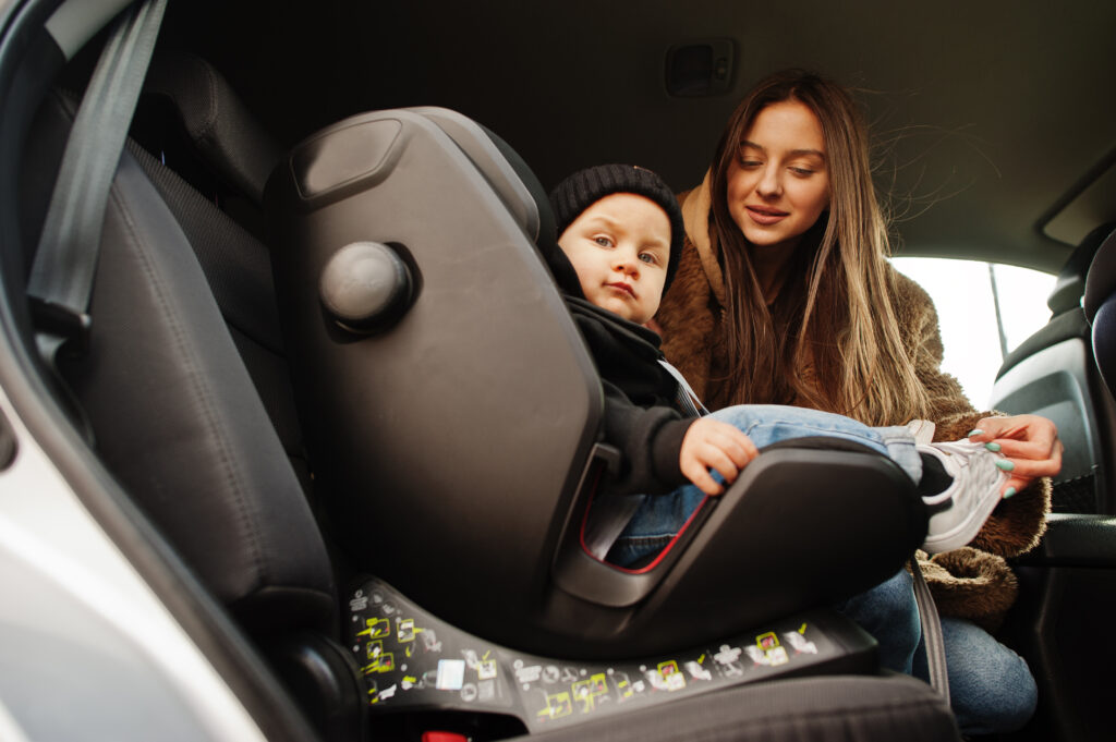 Madre che sistema il figlio nel seggiolino auto i-Size installato correttamente sul sedile posteriore, a testimonianza delle norme di sicurezza 2024 e dell’obbligo del dispositivo anti-abbandono per i bambini fino a 4 anni.