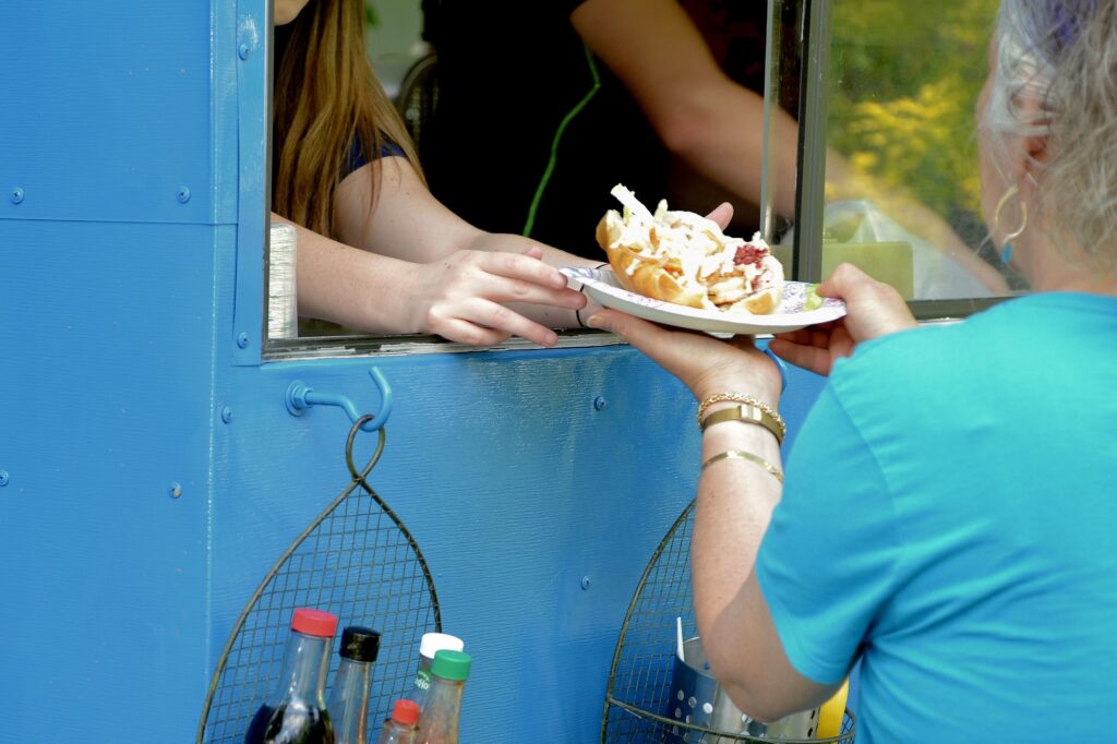 Una donna di spalle prende al bancone di un food truck un piatto con un panino ricolmo di patatine, carne e maionese: si vede il braccio sinistro della cliente e si vedono entrambe le mani della venditrice che sporgono dal finestrino del furgoncino blu